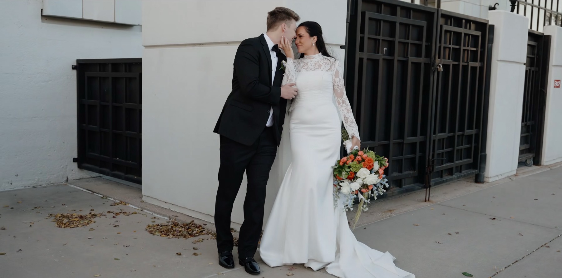 Bride and groom leaning together by a black iron gate, her lace sleeves and an orange bouquet