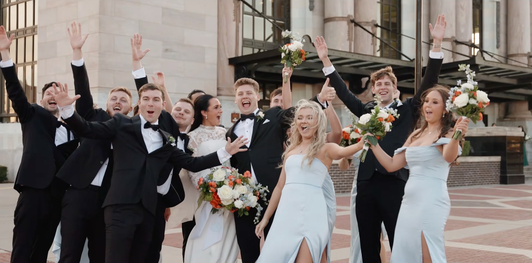 The whole wedding party cheering with their arms raised outside a stone building