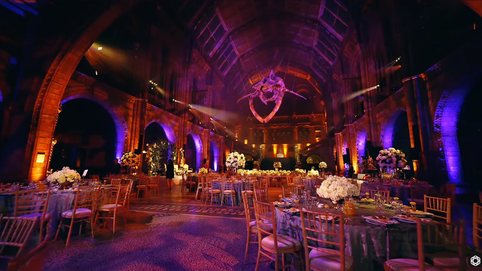 A vaulted stone hall lit in purple and amber, round tables laid and flowers set for the reception