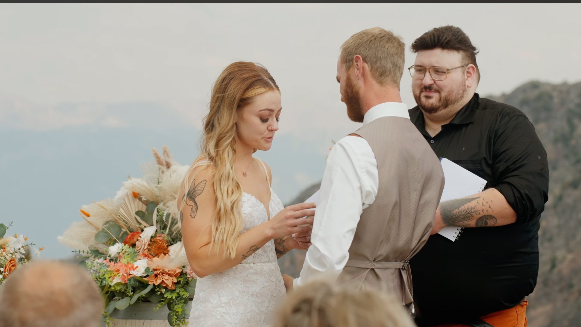 Bride and groom facing a celebrant during an outdoor mountain ceremony, dried flowers in her bouquet