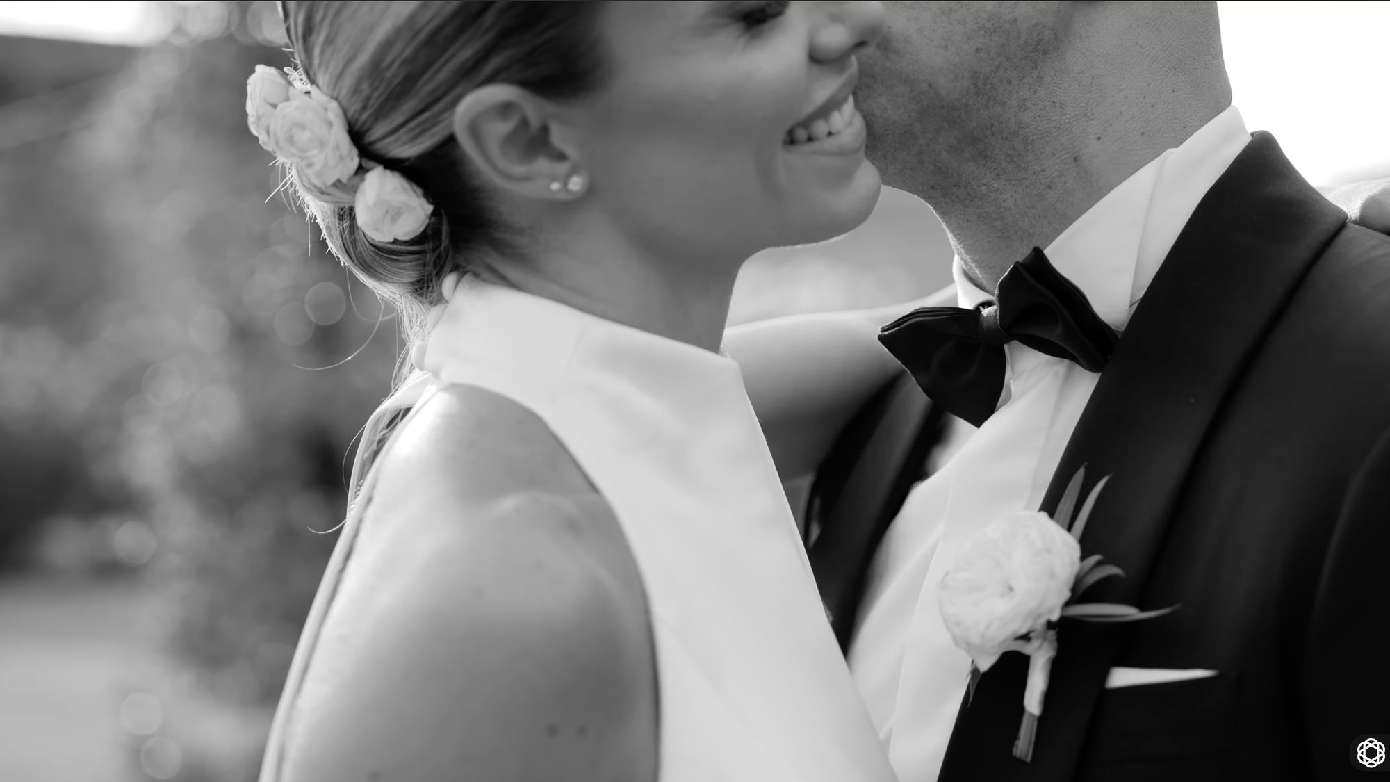 Black and white portrait of a bride resting against the groom’s shoulder, his bow tie and buttonhole in frame