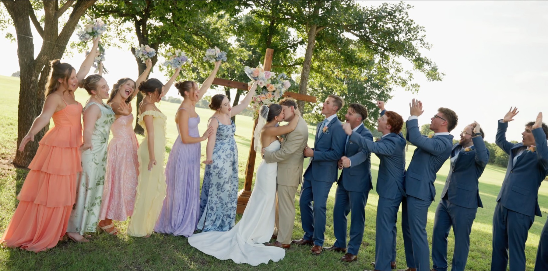 The wedding party cheering in pastel dresses and blue suits as the couple kiss beneath the arch
